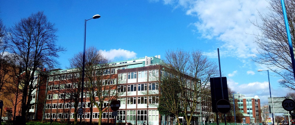 A multi-story red brick and glass office or academic building under a blue sky with scattered clouds, surrounded by leafless trees and street signs.