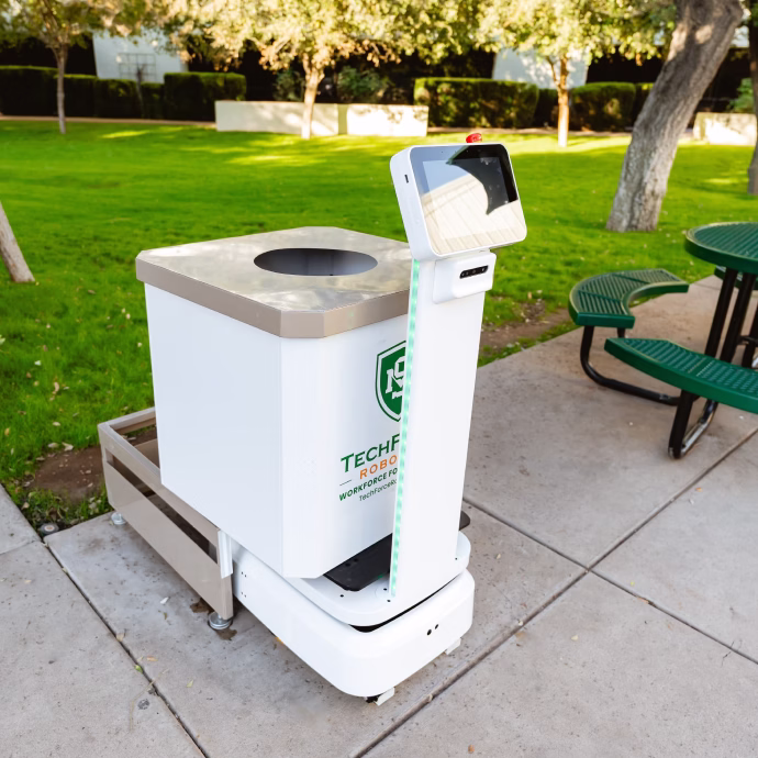 White autonomous robot with a screen mounted on a pole, next to a park bench and green grass area.
