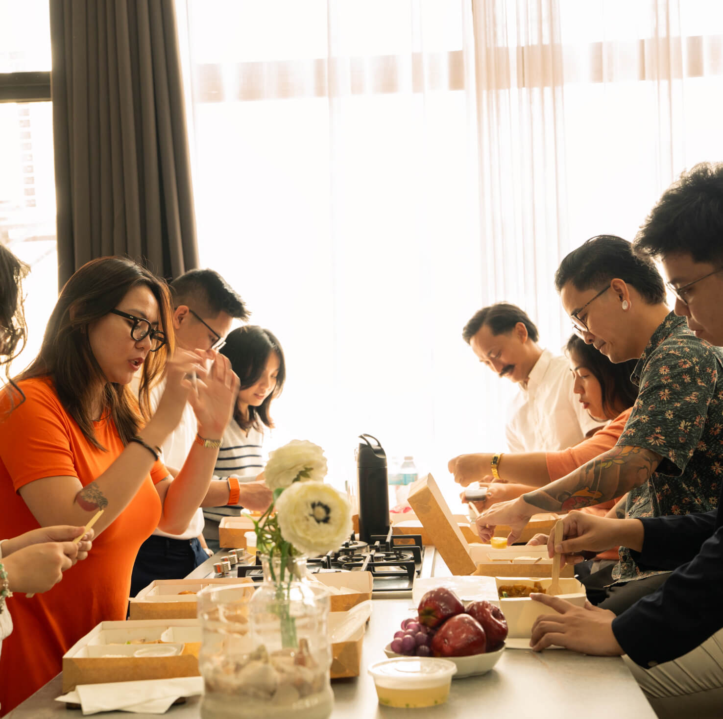 Group of people sitting at a table eating takeaway food together in a bright room with sheer curtains.
