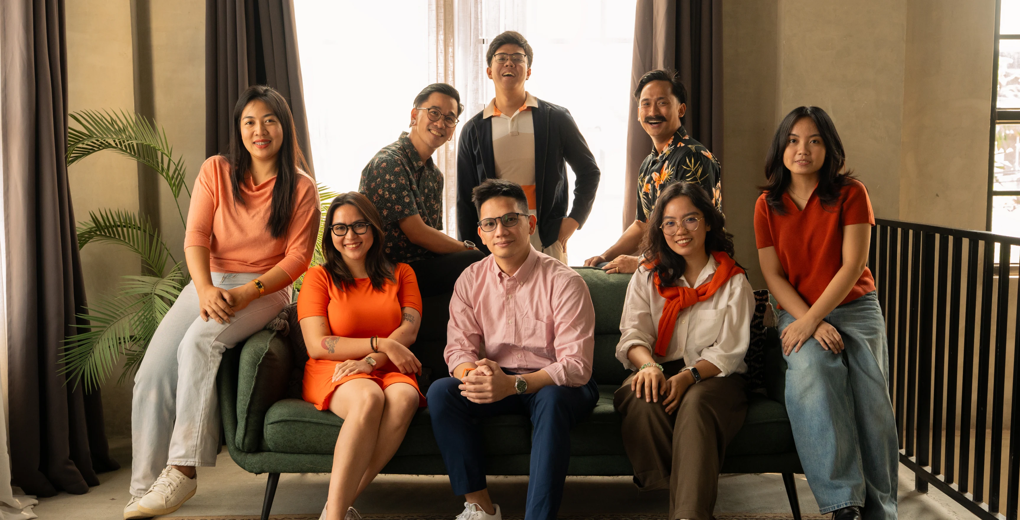 Group of eight diverse young adults sitting and standing around a green couch in a warmly lit room.