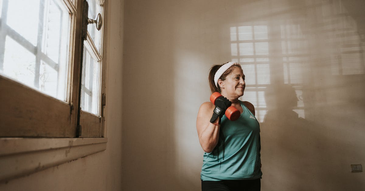 Senior woman in a brightly lit room exercises with dumbbells, promoting fitness at home