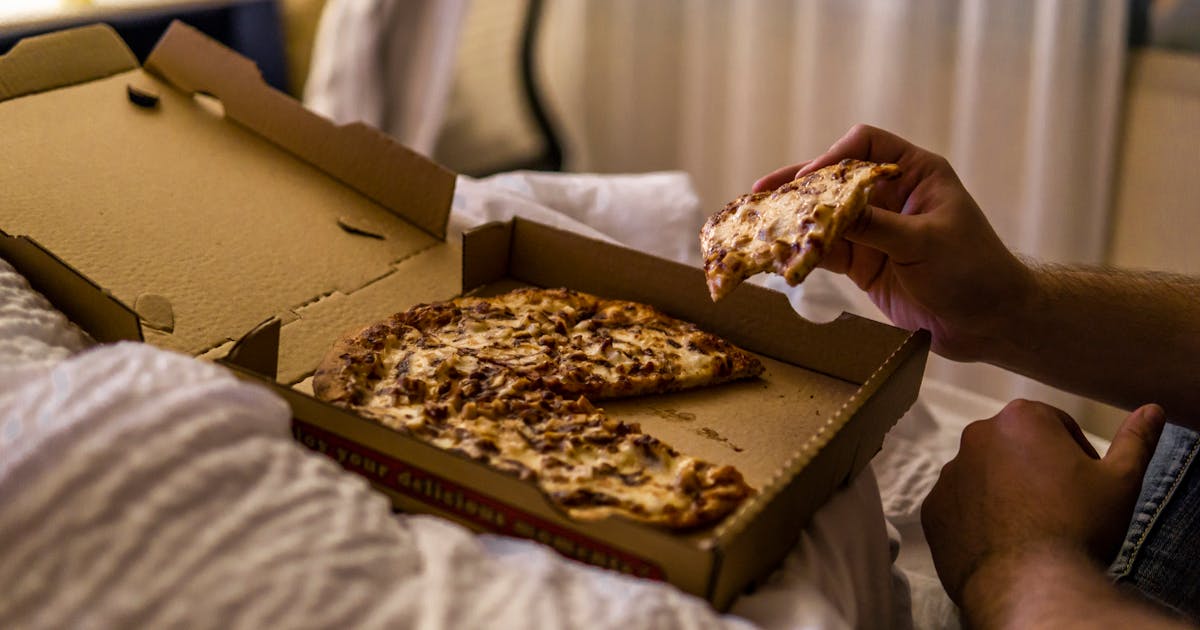 From above side view of person eating tasty slice of pizza with cheese in takeaway cardboard box in bed in apartment