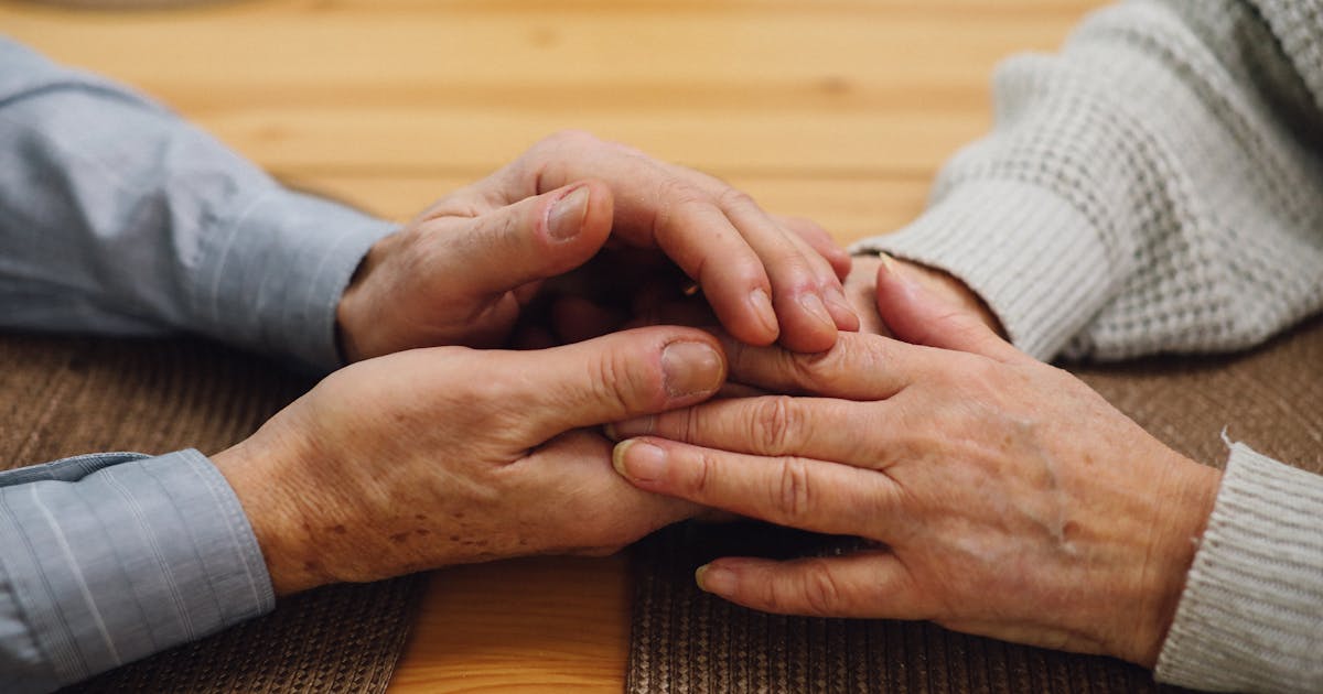 Close-up of an elderly couple holding hands on a wooden table, signifying support