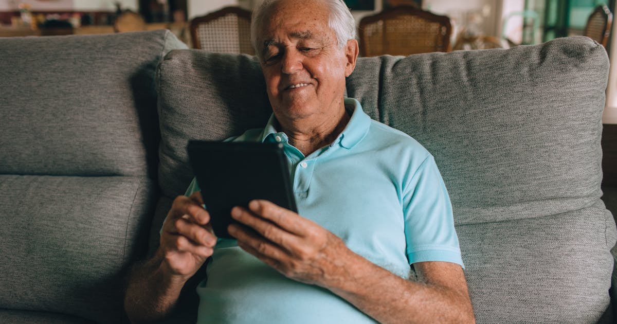 Elderly man in blue polo using a tablet on a sofa, smiling indoors