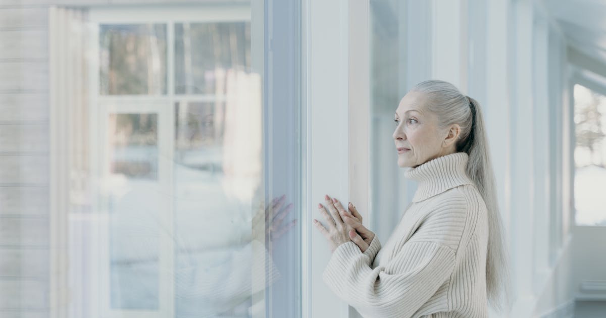 Serene elderly woman with grey hair looking out a window in a bright room wearing a cozy sweater