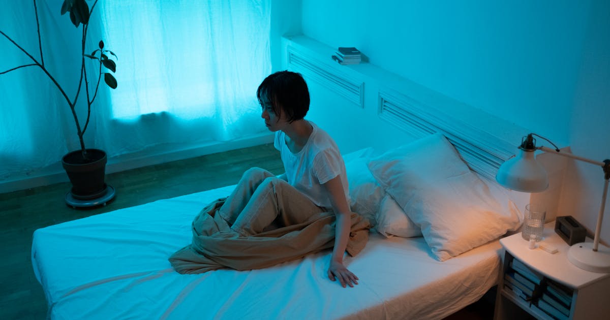Woman sitting on bed in dimly lit room conveying themes of insomnia and contemplation