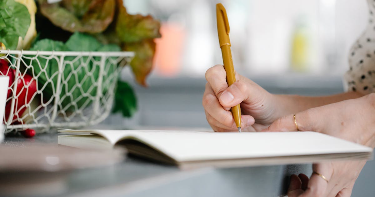 Woman writing in a notebook on a kitchen counter beside a basket of fresh vegetables