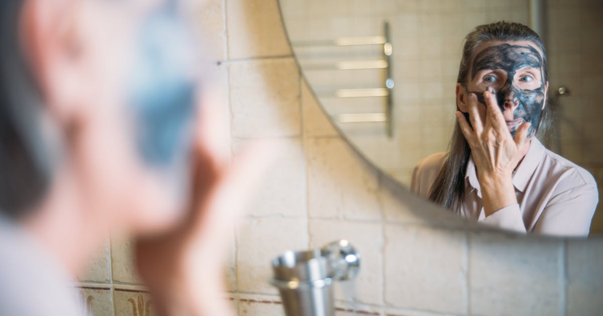 An elderly woman applying a face mask while looking in the bathroom mirror promoting self-care