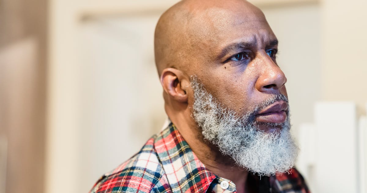 A thoughtful portrait of a bald, bearded man in a plaid shirt, looking away indoors