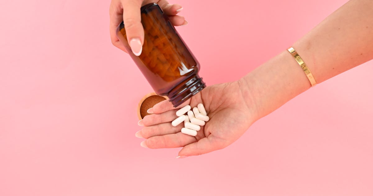 Hands pouring white supplement capsules from a brown bottle on a bright background