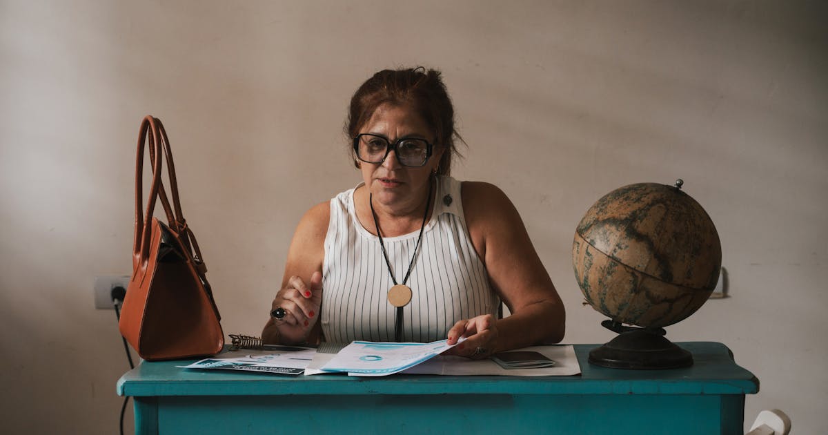 A middle-aged woman sits at her home desk with documents, a globe, and a bag