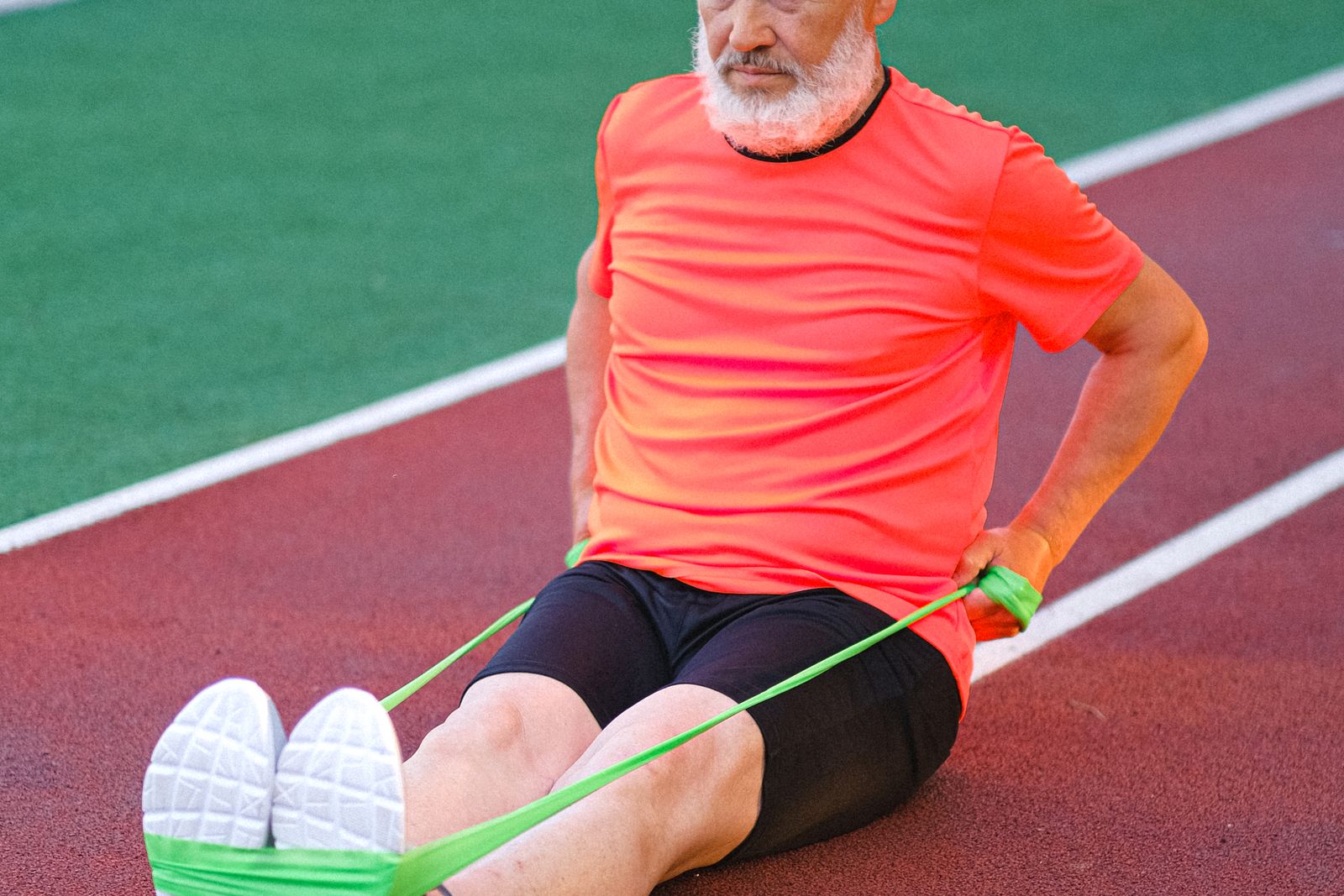 An older man seated on a running track pulling a green resistance band with both hands