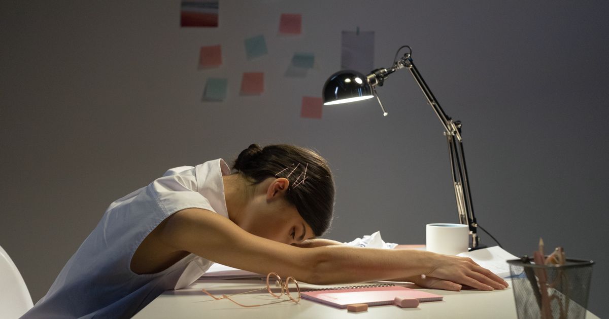 An exhausted woman resting her head on her arms at a desk in the evening light