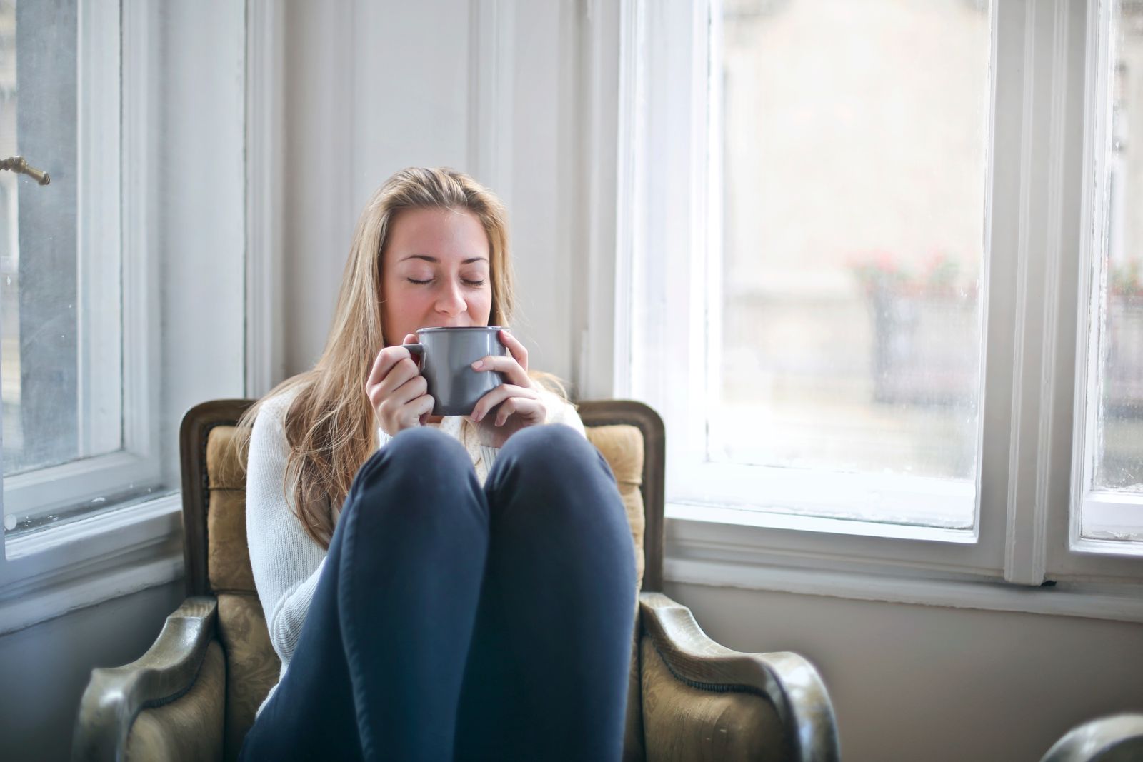 A woman sitting in an armchair by a window with her eyes closed, holding a mug of tea