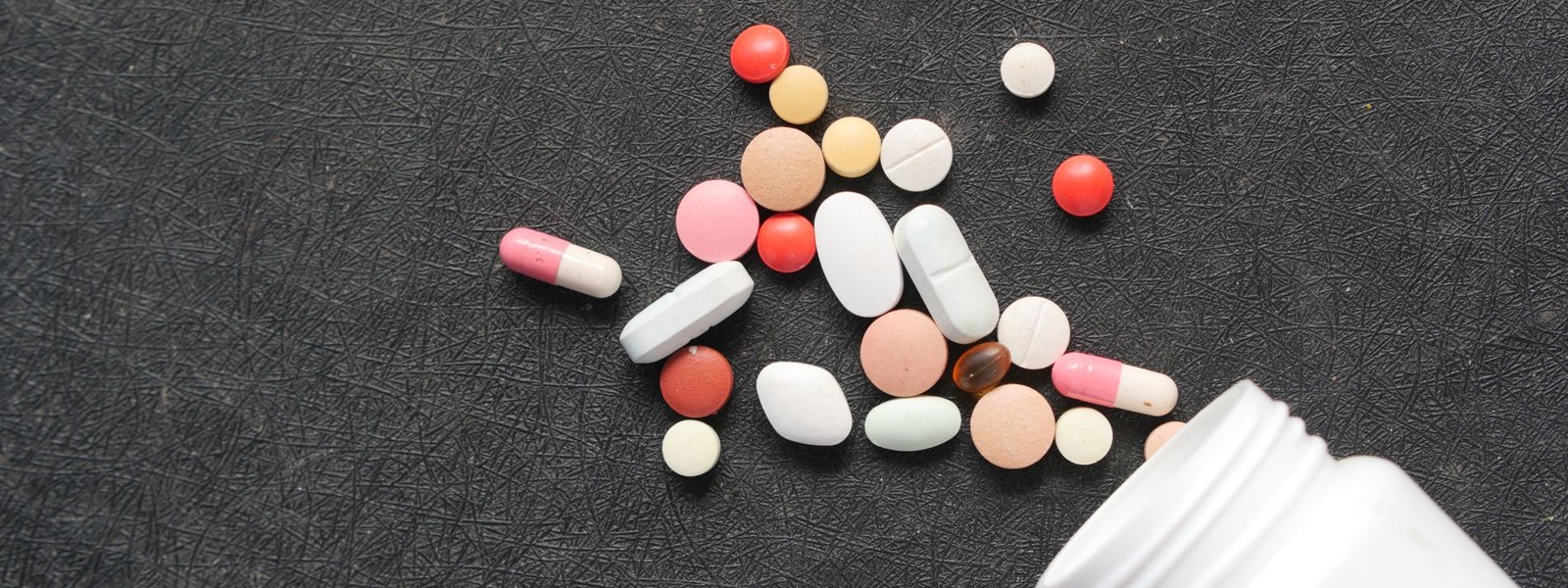 An assortment of differently shaped tablets and capsules spilling from a white bottle onto a dark surface