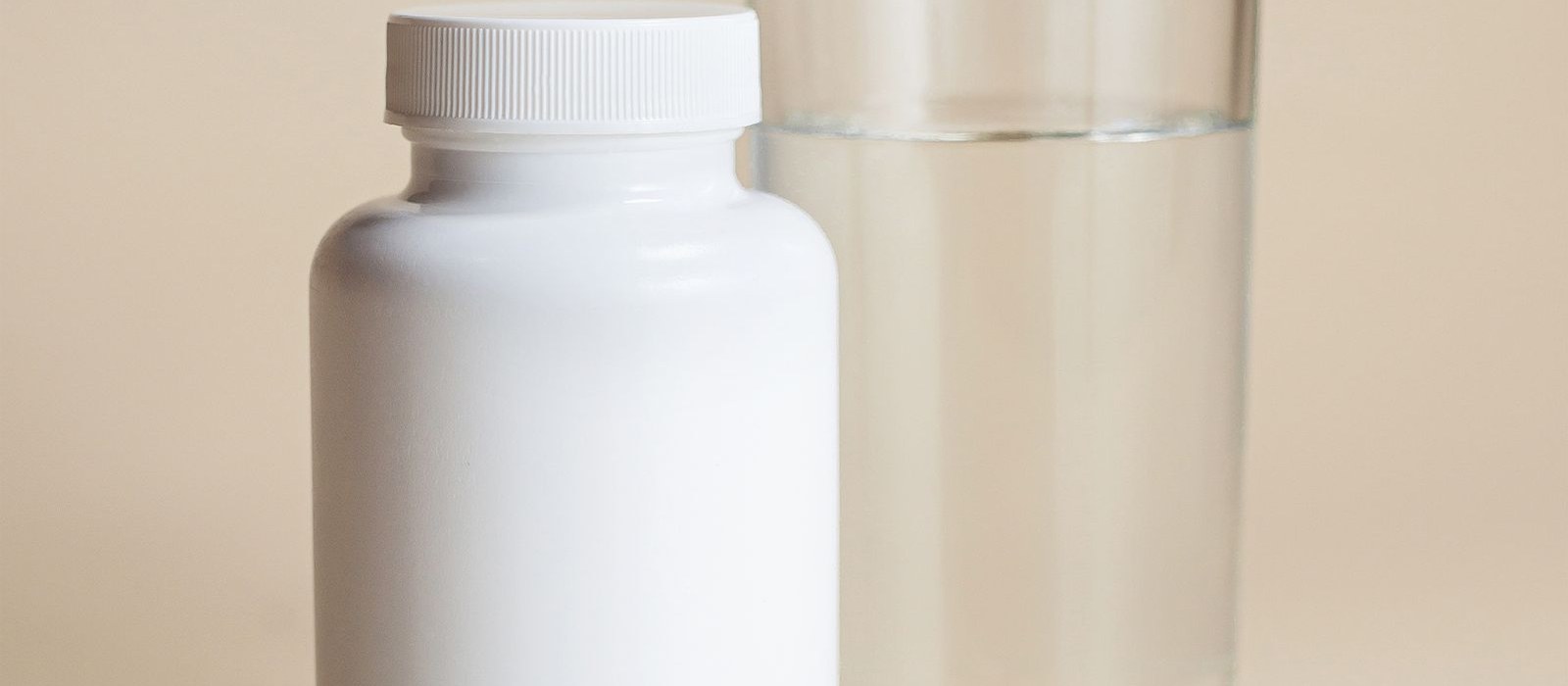 A plain white supplement bottle standing beside a clear glass of water on a warm neutral surface