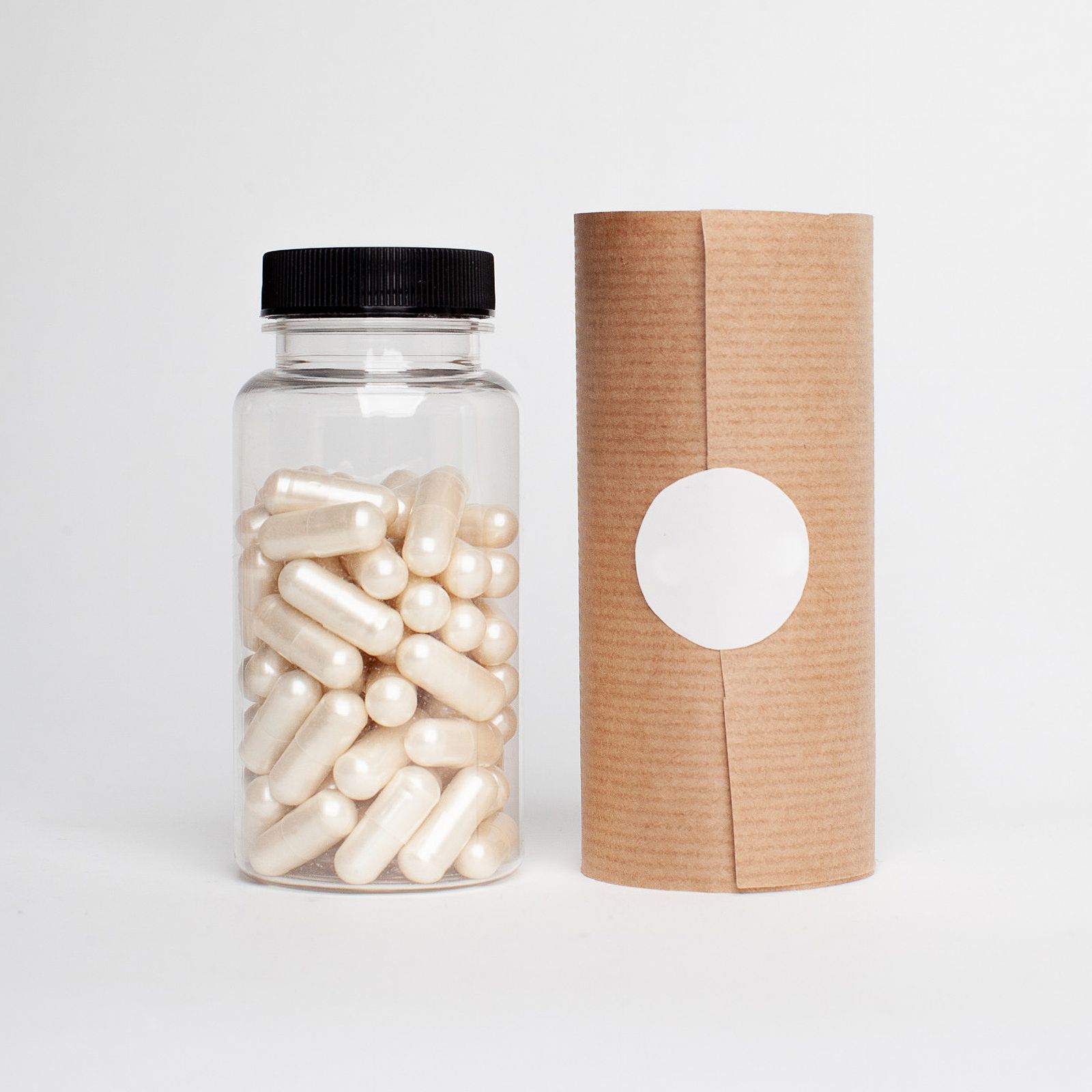 A clear unlabelled supplement bottle filled with capsules beside a plain kraft paper tube