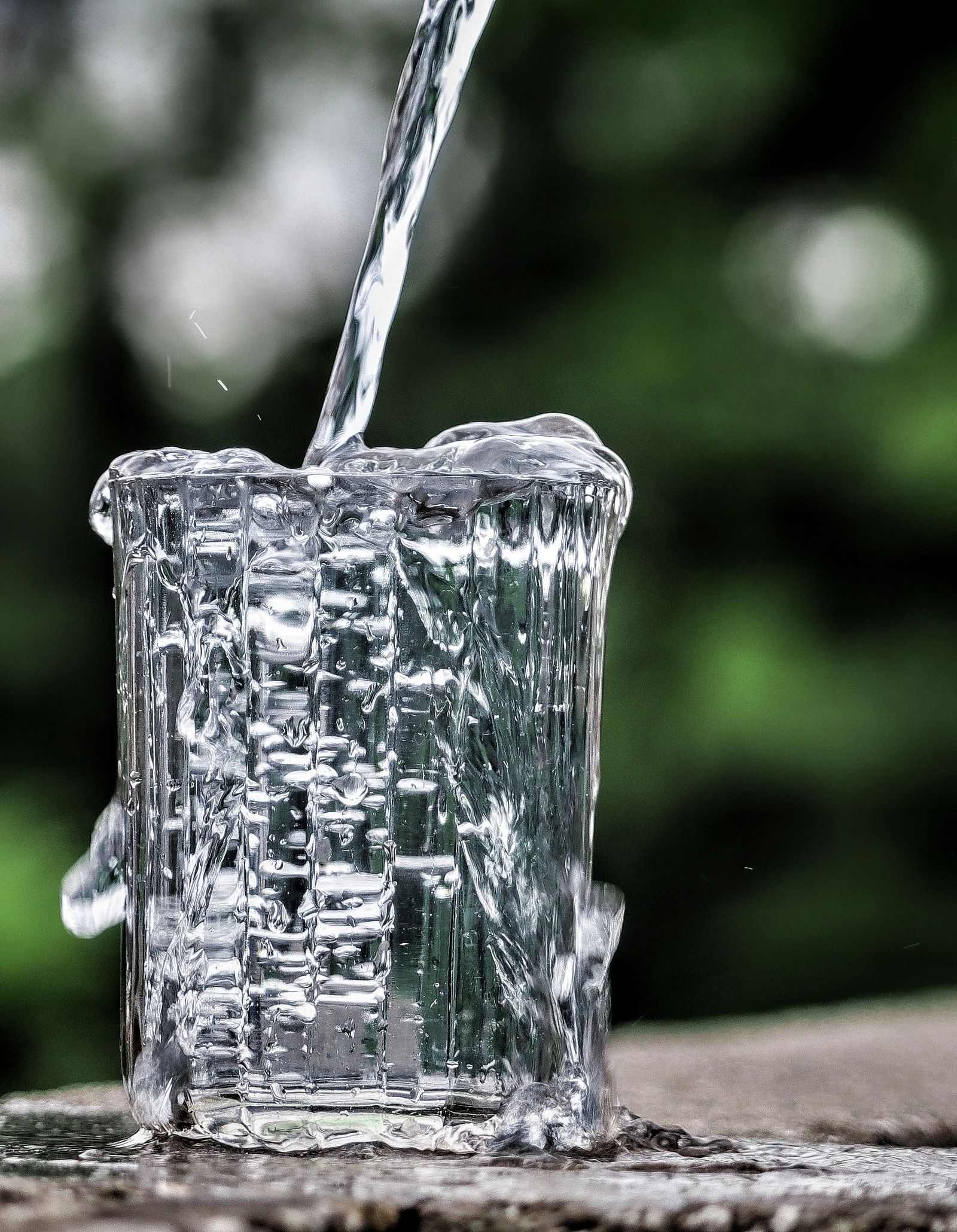 Water pouring into a clear glass until it overflows, on a wooden table outdoors