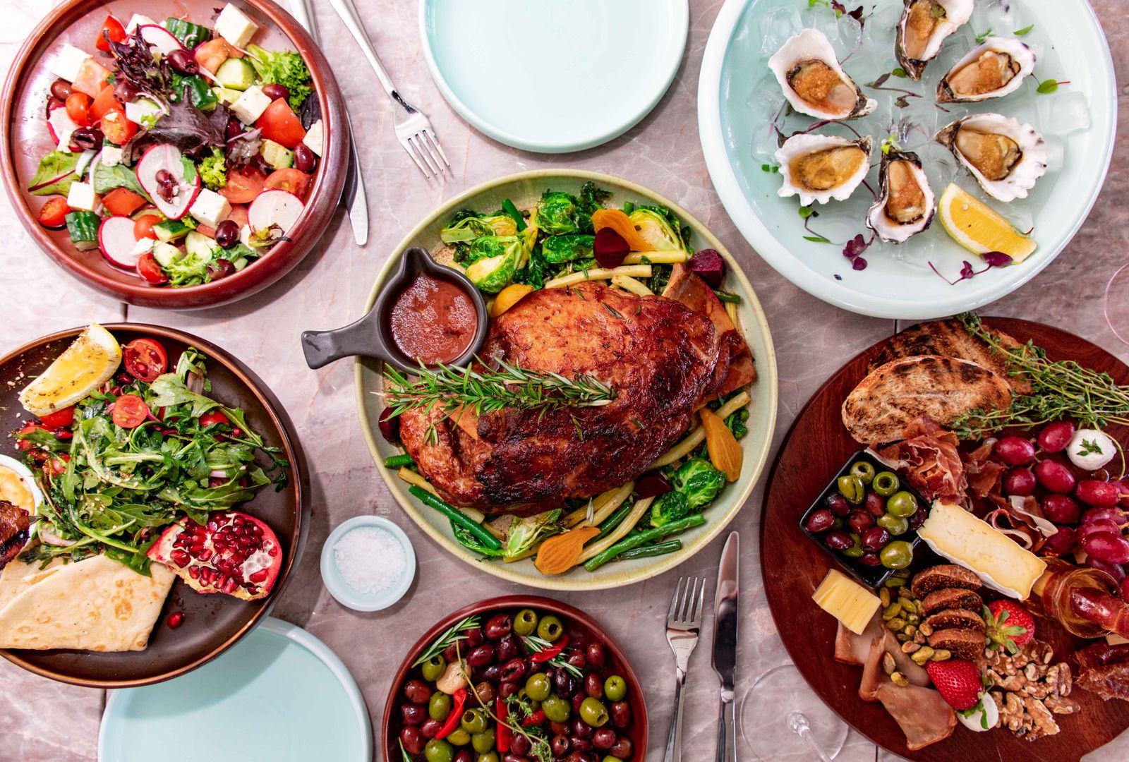 An overhead view of a shared table set with roast meat, salads, olives, oysters and a cheese board