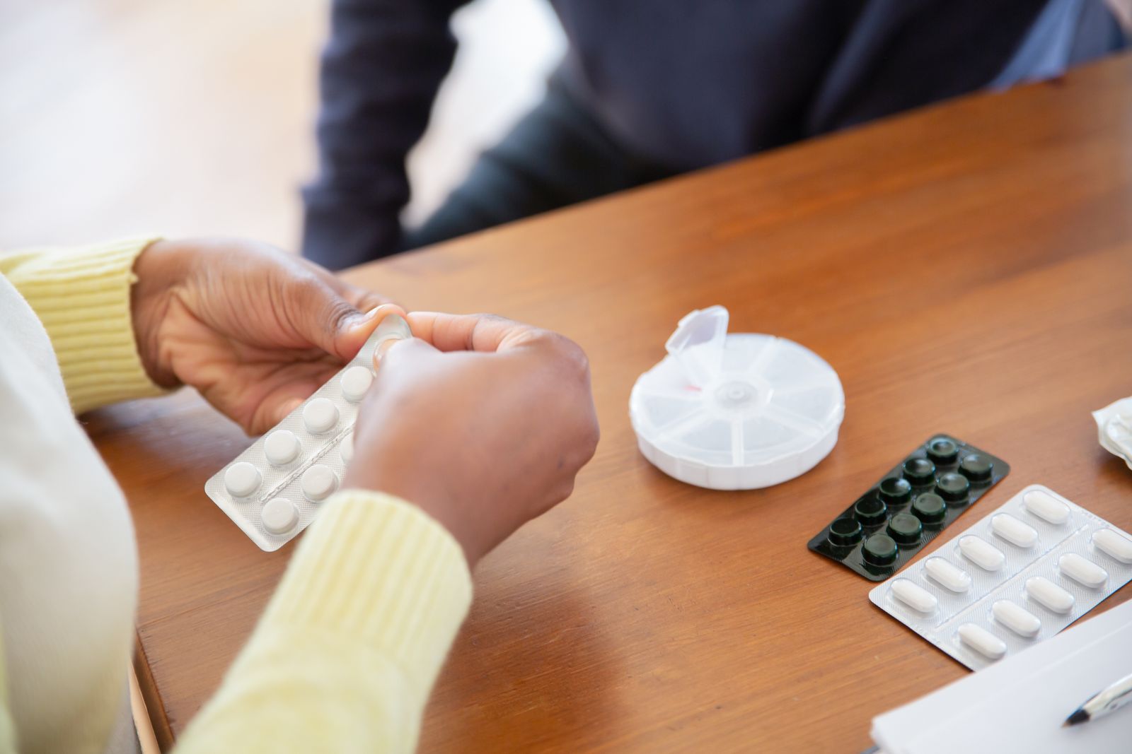 Hands sorting tablets from blister packs beside a weekly pill organiser on a wooden table