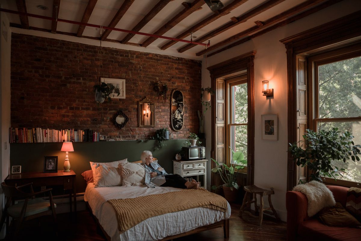A woman resting on a bed in a warm bedroom, looking out toward a sunlit window
