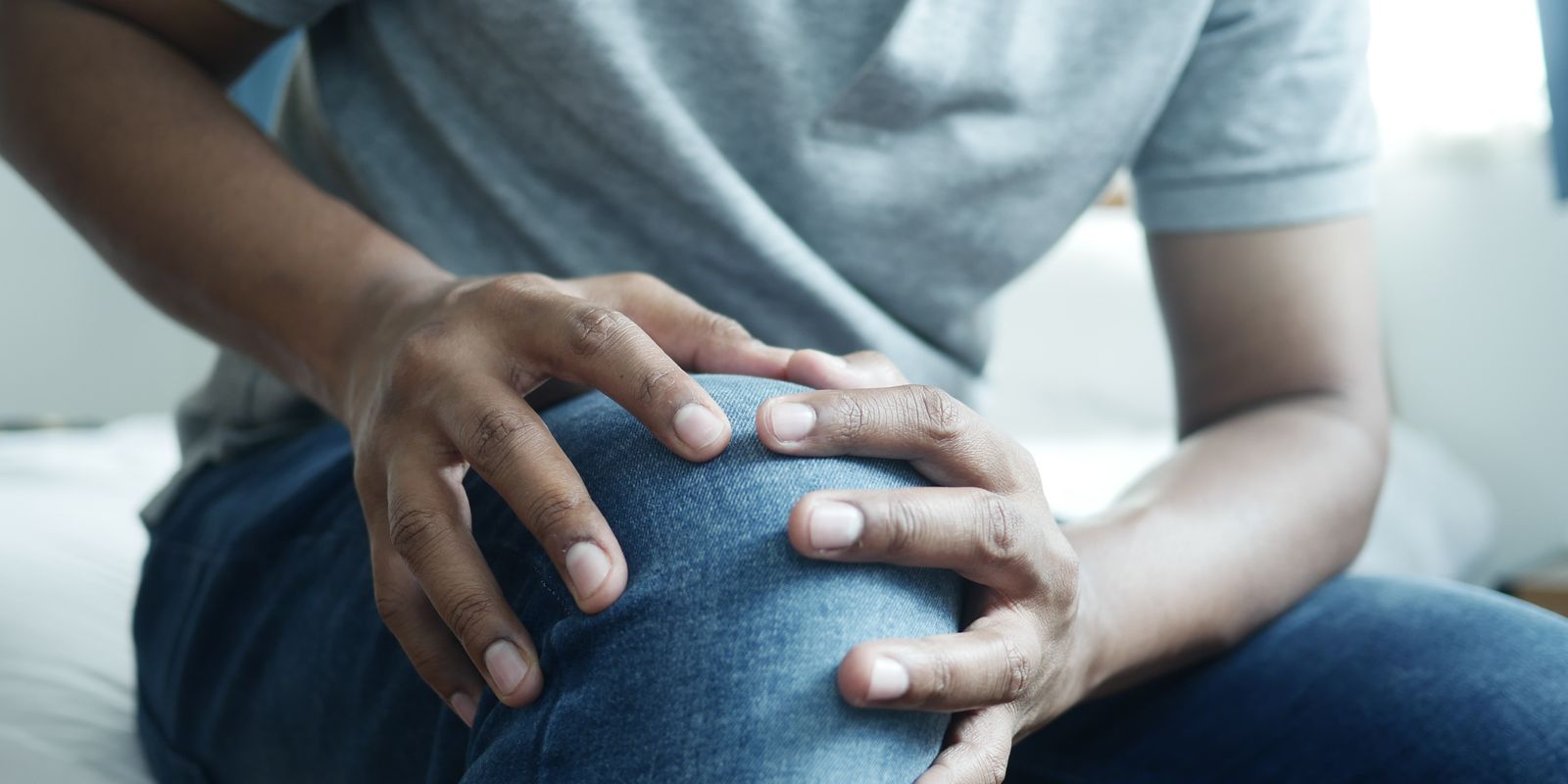 A man sitting on a bed holding his painful knee with both hands