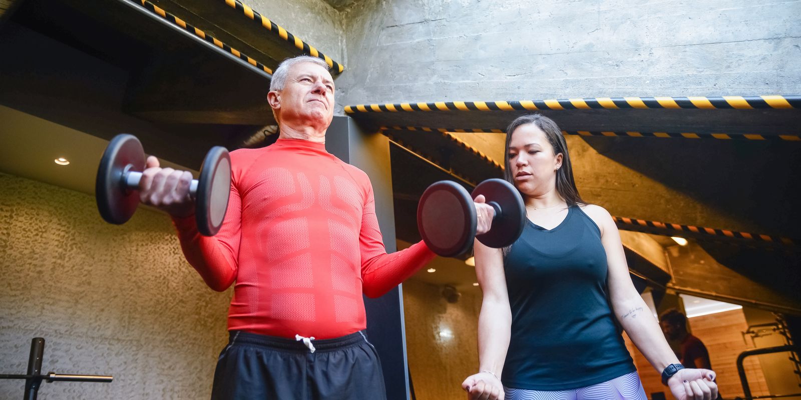 Senior man doing dumbbell curls in a gym beside a trainer