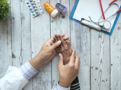 A doctor holds an older patient's hands across a desk with medications nearby