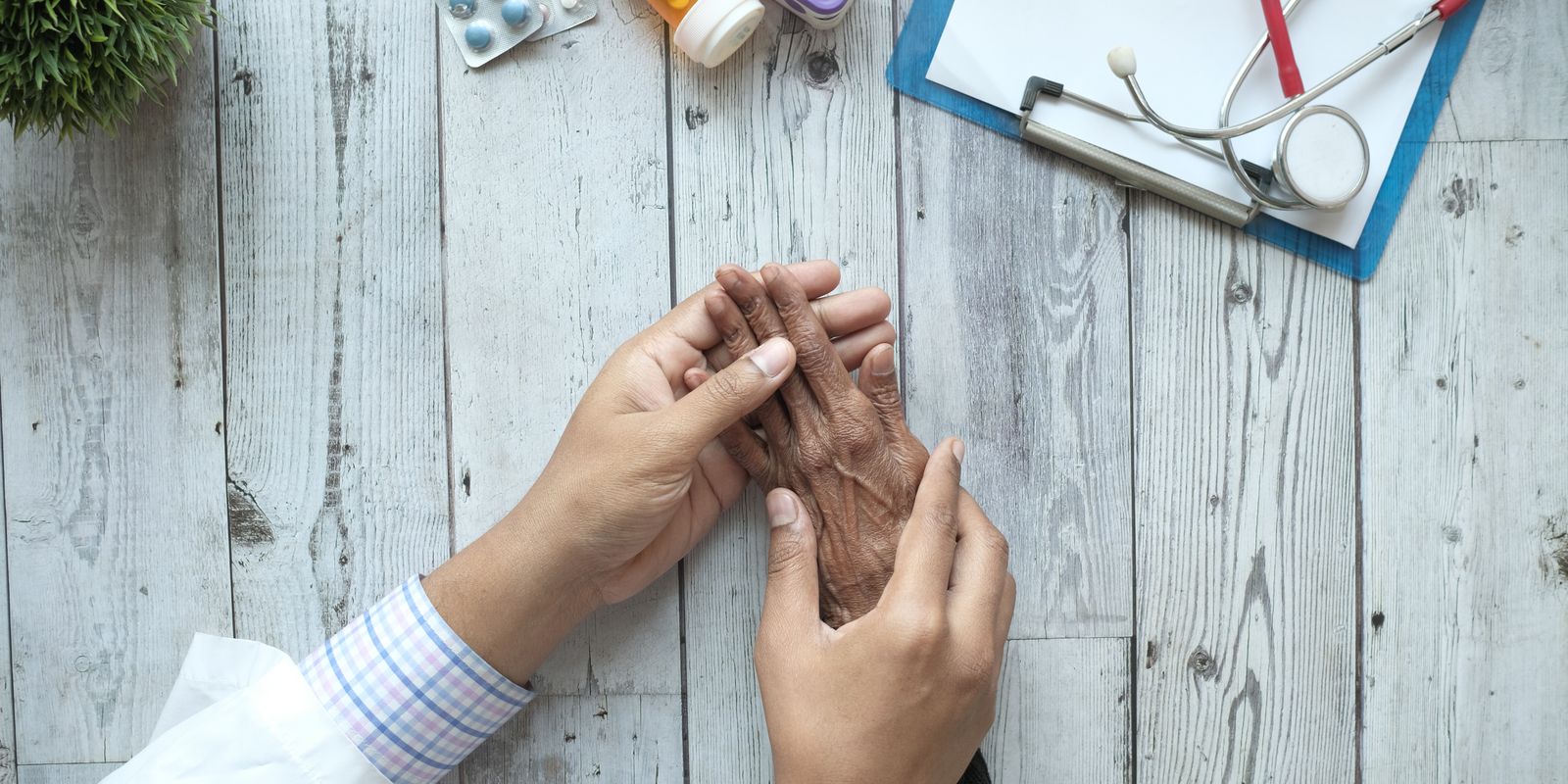 A doctor holds an older patient's hands across a desk with medications nearby