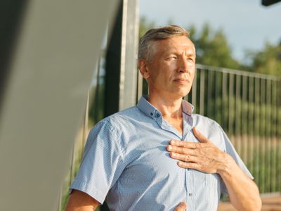 A mature man outdoors rests his hand on his chest with a calm expression