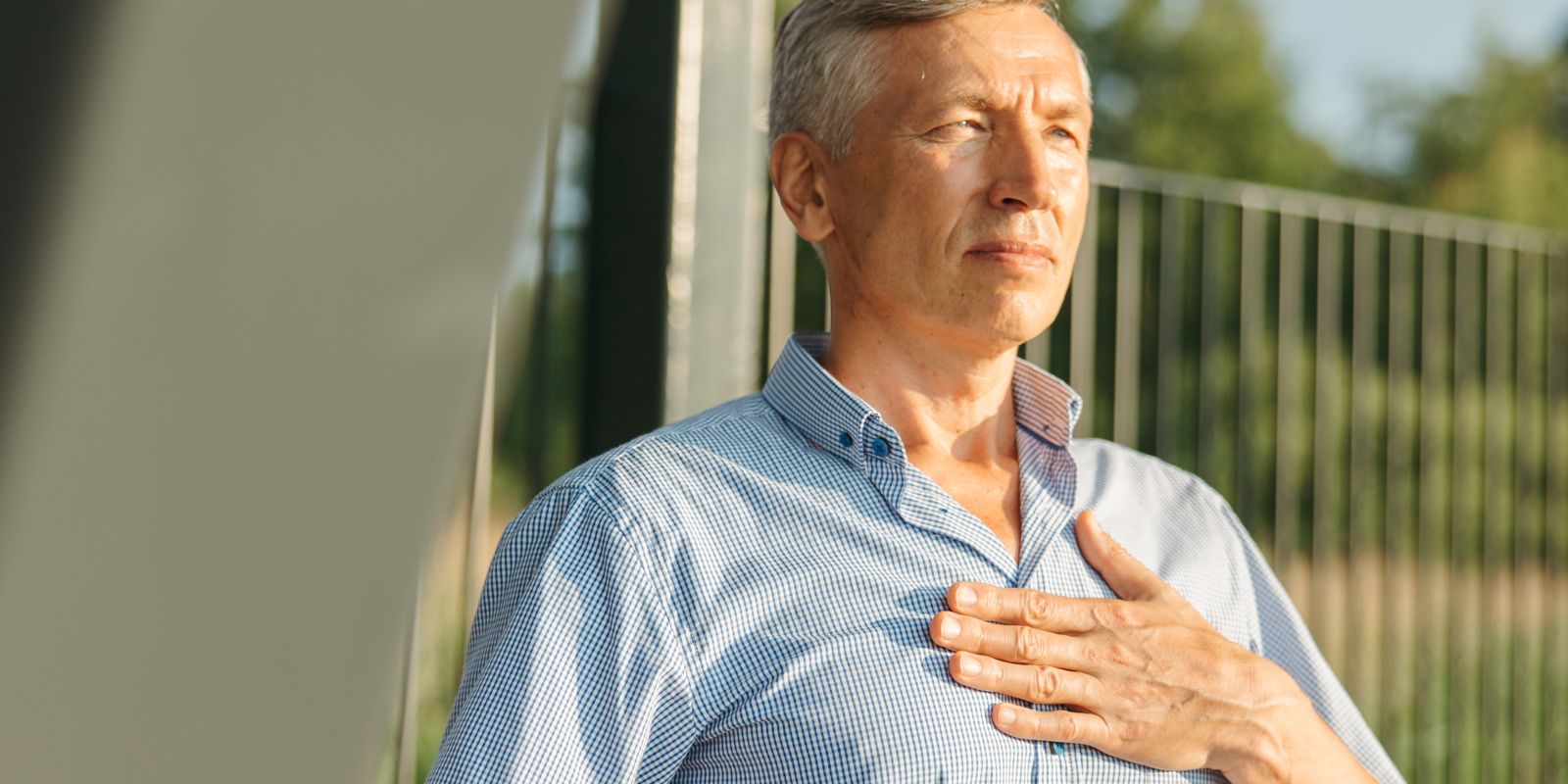 A mature man outdoors rests his hand on his chest with a calm expression