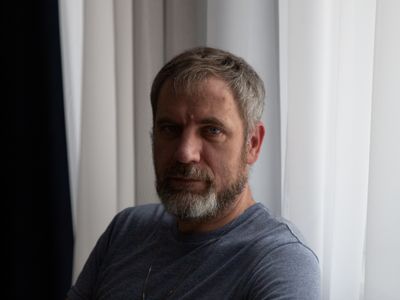 A middle-aged man with a gray beard stands by a window in soft light