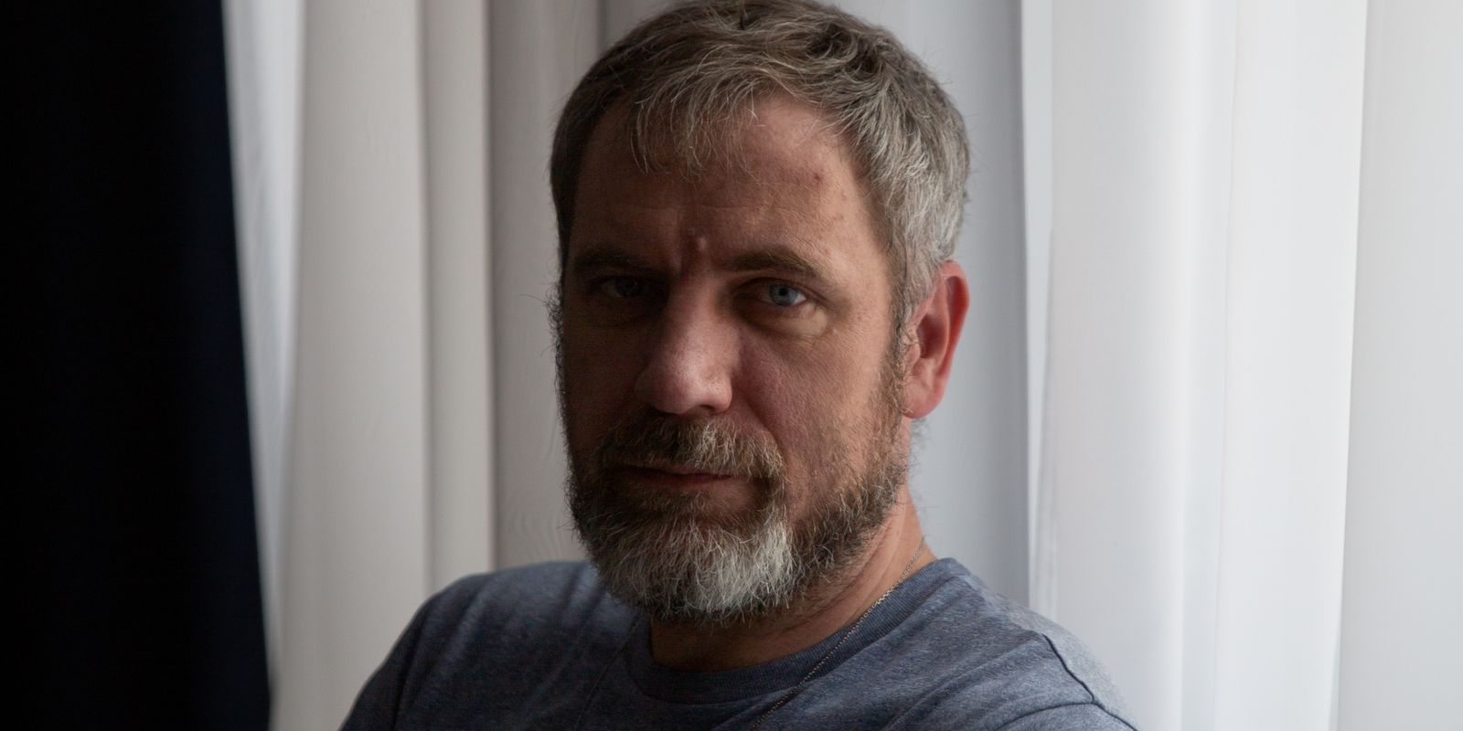 A middle-aged man with a gray beard stands by a window in soft light