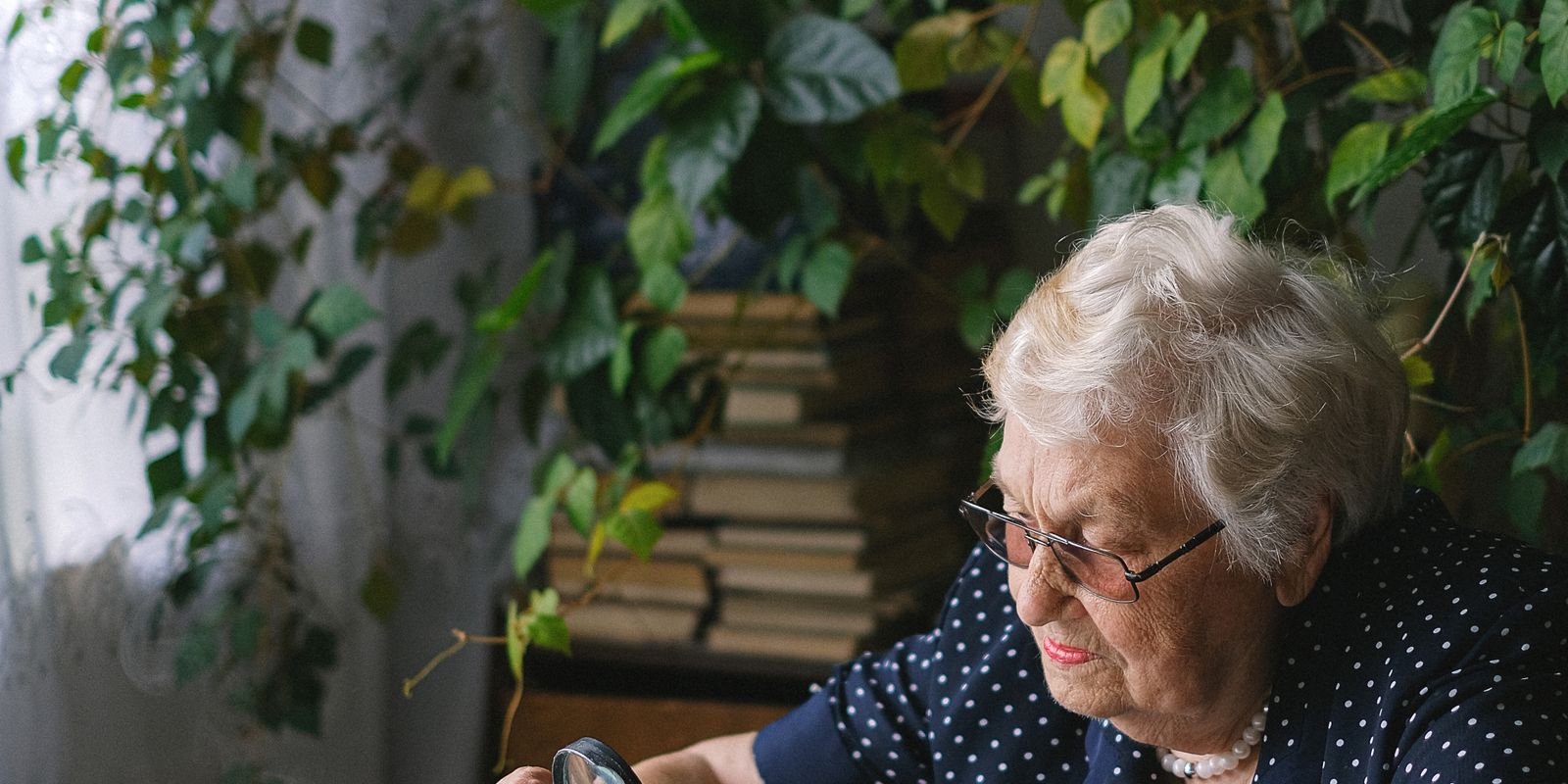 An older woman wearing glasses reads at a table near a window
