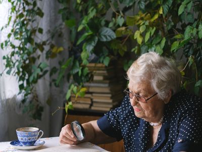 An older woman wearing glasses reads at a table near a window