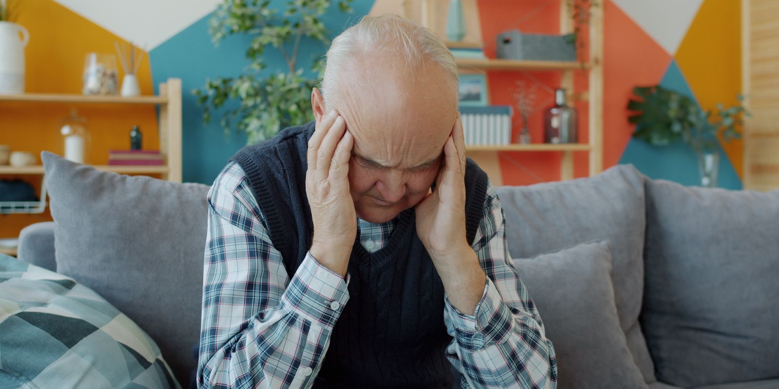 An older man sitting at home with both hands pressed against his head