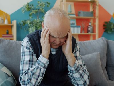 An older man sitting at home with both hands pressed against his head