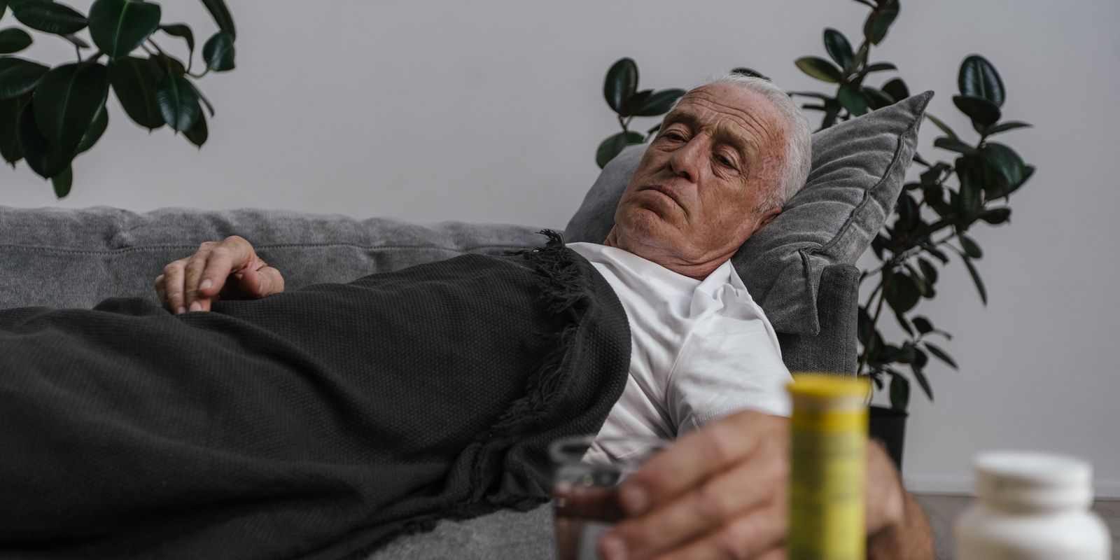 An older man resting on a sofa at home holding a glass of water, with medicine bottles on the table