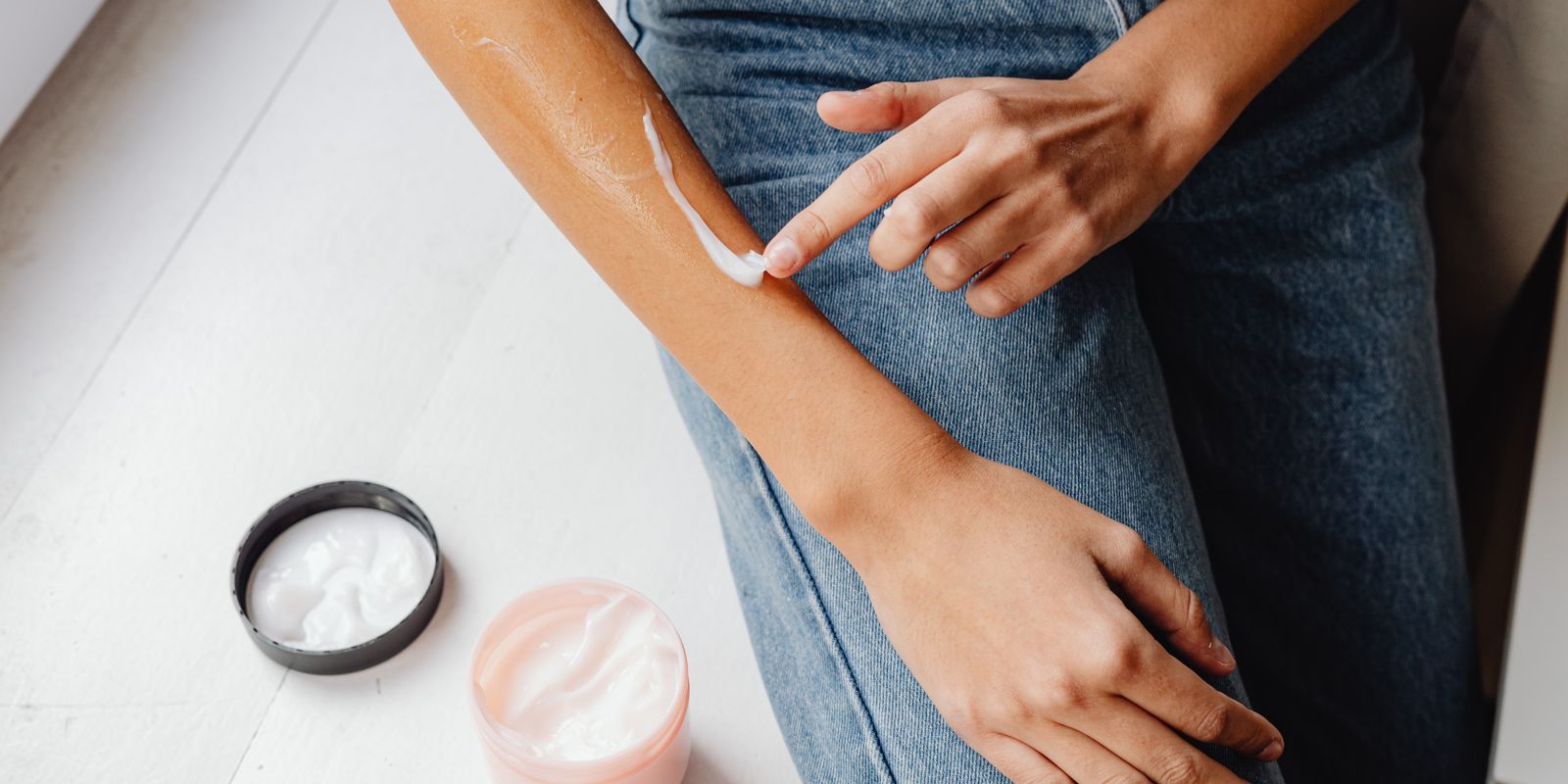 A person spreading a stripe of white cream along a forearm, with two open jars of moisturizer nearby