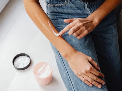 A person spreading a stripe of white cream along a forearm, with two open jars of moisturizer nearby