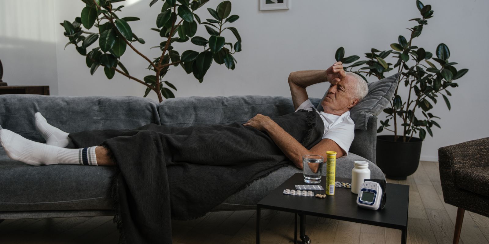 An older man resting on a sofa beside a table holding medication bottles, a blister pack and a blood pressure monitor