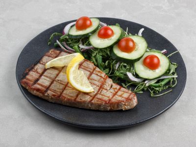 A grilled tuna steak on a dark plate with lemon wedges and a cucumber salad