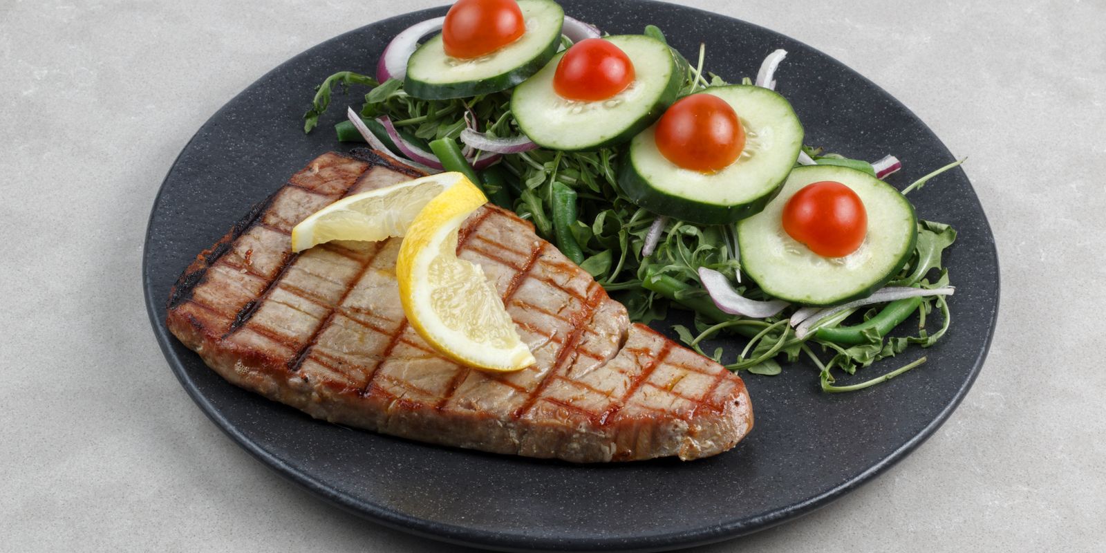A grilled tuna steak on a dark plate with lemon wedges and a cucumber salad