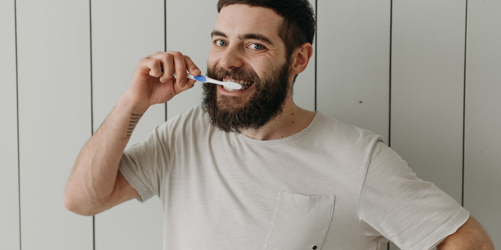A bearded man in a light t-shirt brushing his teeth in a bathroom