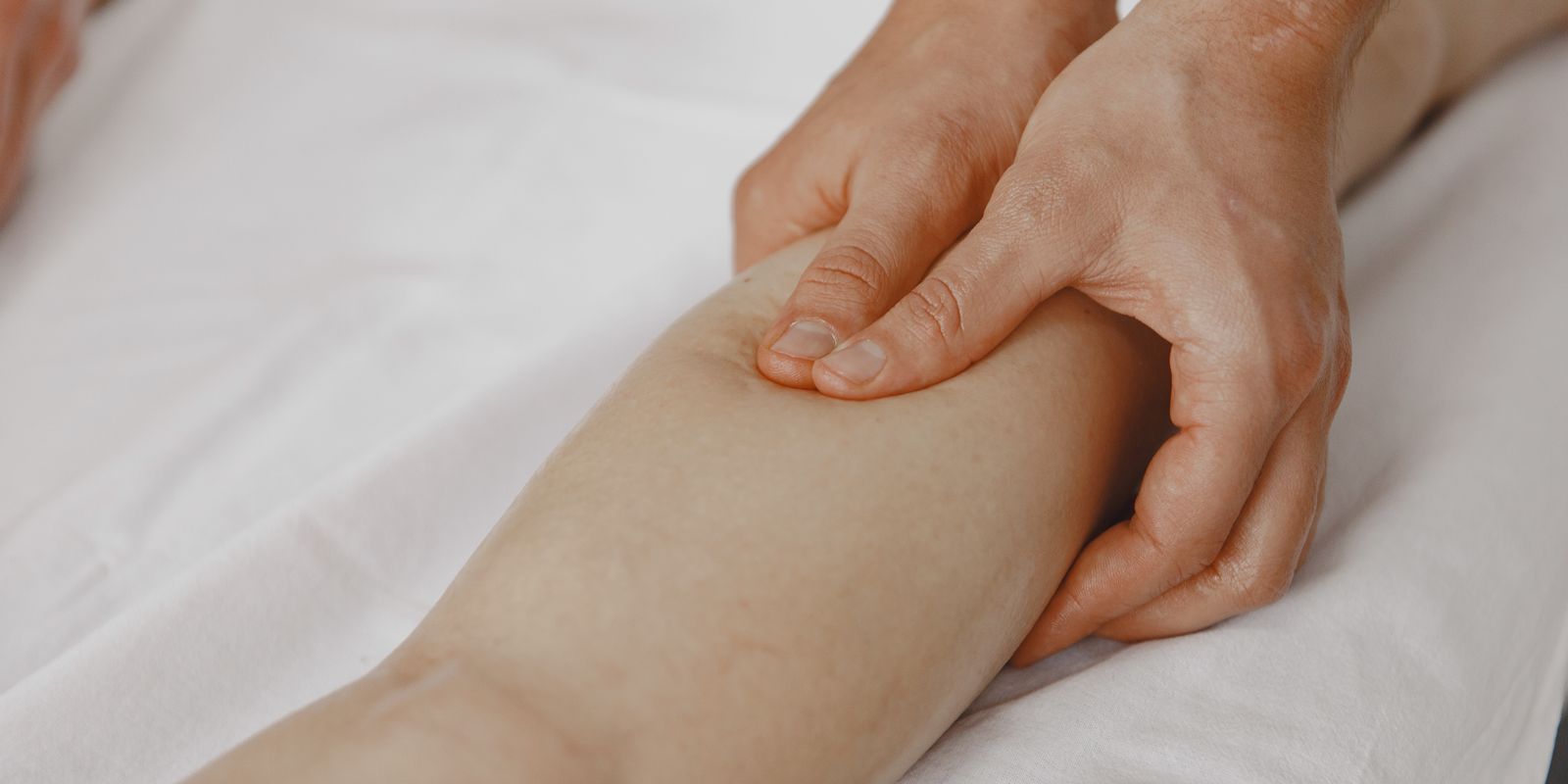 Hands massaging a bare calf on a white bed sheet