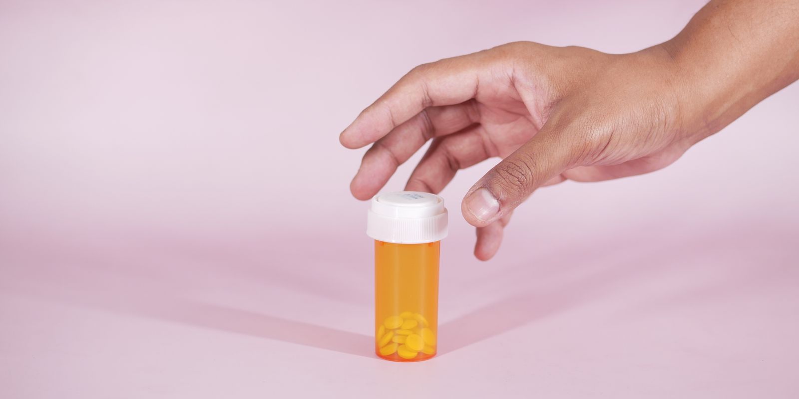 A hand reaching for an amber prescription pill bottle on a pink surface