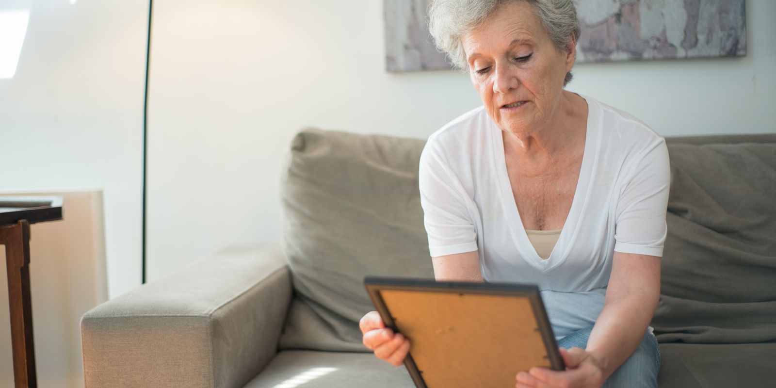 An older woman sitting on a sofa at home looking down at a framed photograph