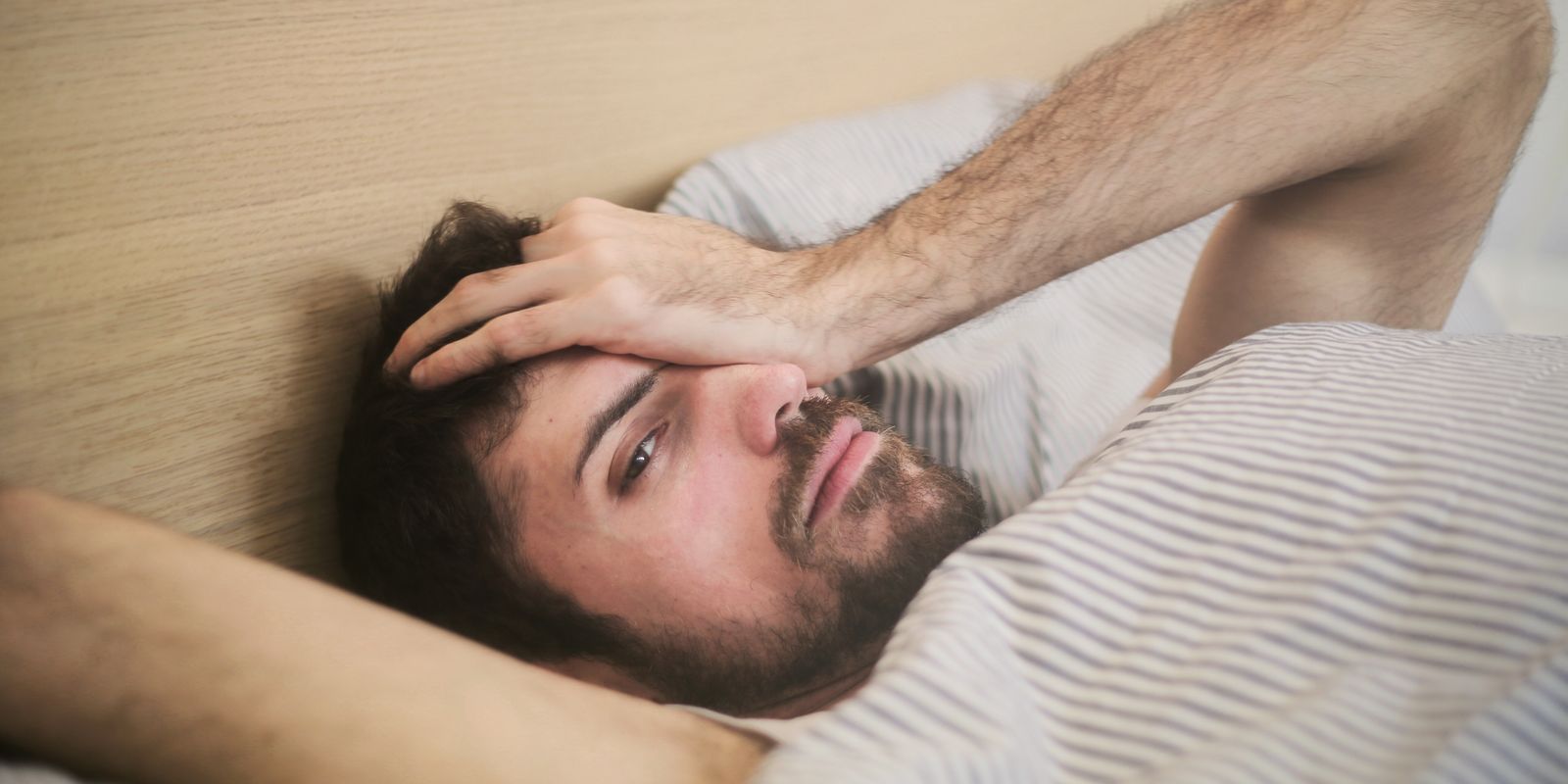 A bearded man lying in bed with his hand pressed to his temple
