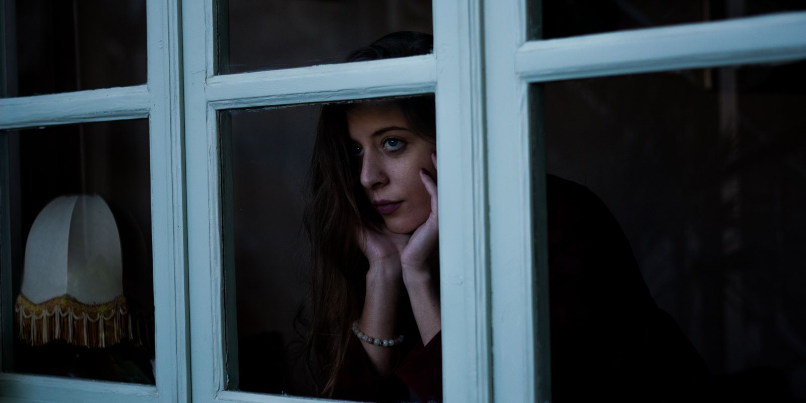 A woman sitting in a dark room looking out through an old window on a grey day