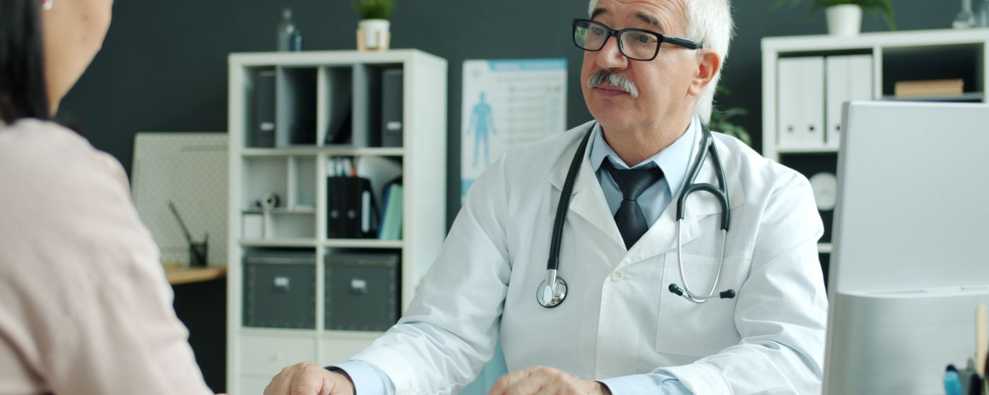 A senior doctor in a white coat explaining something across a desk to a patient